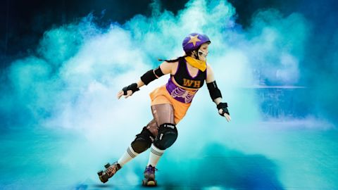 A roller derby athlete in safety gear skates on a lit arena floor, surrounded by blue smoke or mist.