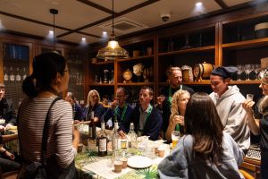 A group of people are seated around a table with drinks and snacks in a warmly lit room, as one person stands and addresses them. Shelves in the background display various bottles and decorative items.