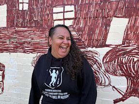 Jinny-Jane Smith stands in front of the Redfern mural 'Mission Boy Dreams' which is based on an etching by Wiradjuri artist Roy Kennedy. It depicts his memories of the Warangesda Mission in the Riverina where his family is from. It’s also the Aboriginal reserve where Jinny-Jane’s grandmother Margaret comes from.


