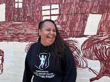 Jinny-Jane Smith stands in front of the Redfern mural 'Mission Boy Dreams' which is based on an etching by Wiradjuri artist Roy Kennedy. It depicts his memories of the Warangesda Mission in the Riverina where his family is from. It’s also the Aboriginal reserve where Jinny-Jane’s grandmother Margaret comes from.


