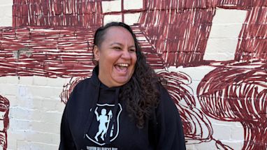Jinny-Jane Smith stands in front of the Redfern mural 'Mission Boy Dreams' which is based on an etching by Wiradjuri artist Roy Kennedy. It depicts his memories of the Warangesda Mission in the Riverina where his family is from. It’s also the Aboriginal reserve where Jinny-Jane’s grandmother Margaret comes from.