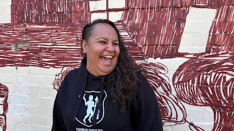 Jinny-Jane Smith stands in front of the Redfern mural 'Mission Boy Dreams' which is based on an etching by Wiradjuri artist Roy Kennedy. It depicts his memories of the Warangesda Mission in the Riverina where his family is from. It’s also the Aboriginal reserve where Jinny-Jane’s grandmother Margaret comes from.