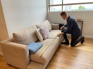 A person kneels next to a beige couch, using a measuring tape to measure its length. The couch has two pillows and a folded blanket on it.