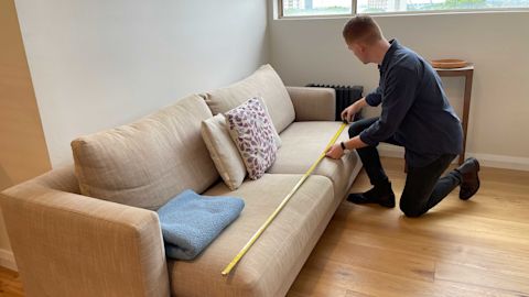 A person kneels next to a beige couch, using a measuring tape to measure its length. The couch has two pillows and a folded blanket on it.