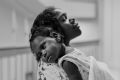 A black and white photo of a person holding a child. The child rests their head on the person's shoulder while looking at the camera. The background is out of focus.