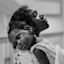 A black and white photo of a person holding a child. The child rests their head on the person's shoulder while looking at the camera. The background is out of focus.