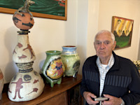 An elderly man stands beside a wooden table displaying several colourful, handmade and hand-painted ceramic vases and pots in a room decorated with paintings.