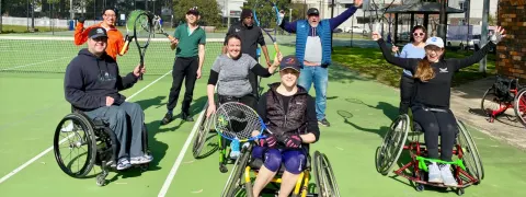 Group shot of adaptive tennis participants; a mixed group of people holding up tennis racquets and their arms for the camera, on a green tennis court. 