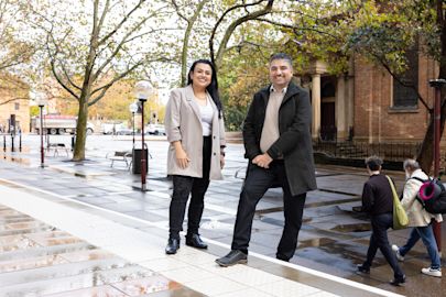 A woman wearing a beige blazer and a man wearing a dark grey coat smile to camera. They standing and leaning in a relaxed way. Trees and street lamps spread out on a wide city pathway behind them.  
