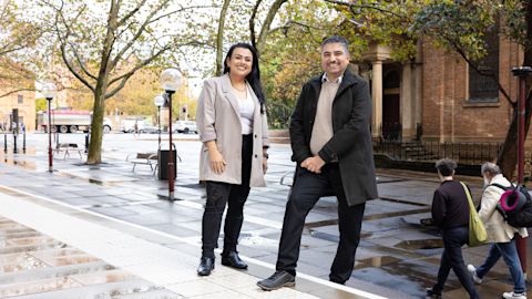 A woman wearing a beige blazer and a man wearing a dark grey coat smile to camera. They standing and leaning in a relaxed way. Trees and street lamps spread out on a wide city pathway behind them.