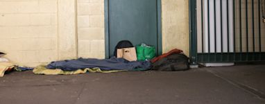 A person sleeping on the street covered with blankets, with belongings nearby.