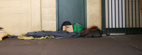 A person sleeping on the street covered with blankets, with belongings nearby.