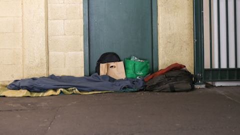 A person sleeping on the street covered with blankets, with belongings nearby.