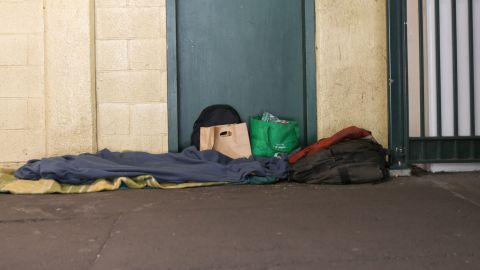 A person sleeping on the street covered with blankets, with belongings nearby.