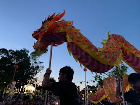 People hold up a colorful dragon puppet during a festival or parade at dusk, with a crowd and trees visible in the background.