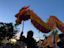 People hold up a colorful dragon puppet during a festival or parade at dusk, with a crowd and trees visible in the background.