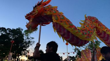 People hold up a colorful dragon puppet during a festival or parade at dusk, with a crowd and trees visible in the background.