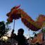 People hold up a colorful dragon puppet during a festival or parade at dusk, with a crowd and trees visible in the background.