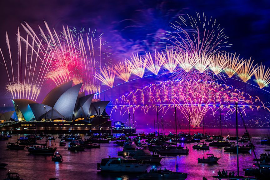 Fireworks illuminate the night sky over the Sydney Opera House and Sydney Harbour Bridge, with boats on the water.