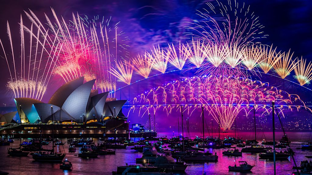 Fireworks illuminate the night sky over the Sydney Opera House and Sydney Harbour Bridge, with boats on the water.