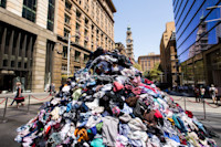 A large pile of mixed clothing sits on an urban street, surrounded by buildings. People walk nearby, and a historic clock tower is visible in the background.