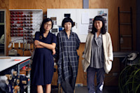 Three women stand in a workspace with sewing supplies, thread spools, and inspiration boards visible in the background.