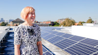 Person with short hair standing on a rooftop with solar panels, smiling. Urban skyline and blue sky in the background.