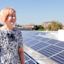 A person with short hair standing on a rooftop with solar panels, smiling. Urban skyline and blue sky in the background.