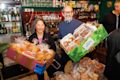 Two people stand in a cafe holding boxes filled with packaged bread and baked goods, smiling at the camera. Shelves with drinks and kitchen equipment are visible in the background.