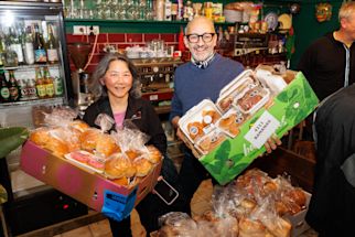 Two people stand in a cafe holding boxes filled with packaged bread and baked goods, smiling at the camera. Shelves with drinks and kitchen equipment are visible in the background.