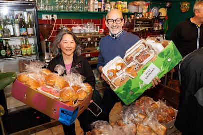 Two people stand in a cafe holding boxes filled with packaged bread and baked goods, smiling at the camera. Shelves with drinks and kitchen equipment are visible in the background.