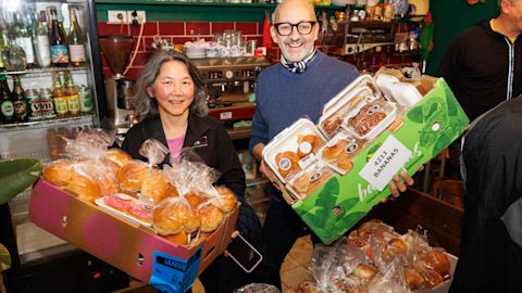 Two people stand in a cafe holding boxes filled with packaged bread and baked goods, smiling at the camera. Shelves with drinks and kitchen equipment are visible in the background.