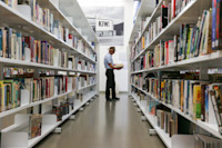 A man in a hat stands in a library aisle, holding a book and browsing shelves filled with books.