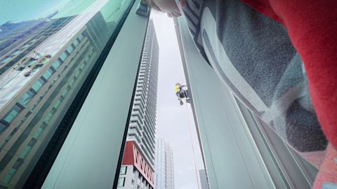 View from inside a building of a window cleaner suspended outside a tall building, seen through glass. Reflections of nearby buildings are visible.