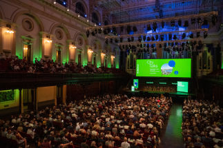 A large audience attends a "City Talks" event in an ornate hall with green and blue lighting. A stage with multiple speakers and large screens displaying event information is visible.