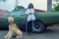 A barefoot child in a white jacket stands with arms crossed, holding a leash attached to a large, shaggy dog. They are in front of a green, rusty, older model car on a gravel surface.