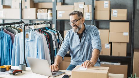 Small business owner wearing glasses and a denim shirt uses his laptop and packs orders on his work desk