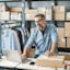 Small business owner wearing glasses and a denim shirt uses his laptop and packs orders on his work desk