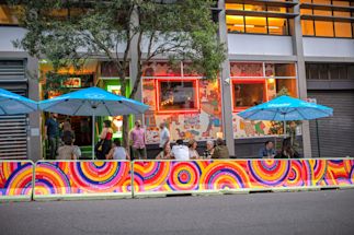 People sit and talk at outdoor tables under blue umbrellas outside a colorful, mural-covered restaurant on a city street.