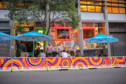 People sit and talk at outdoor tables under blue umbrellas outside a colorful, mural-covered restaurant on a city street.