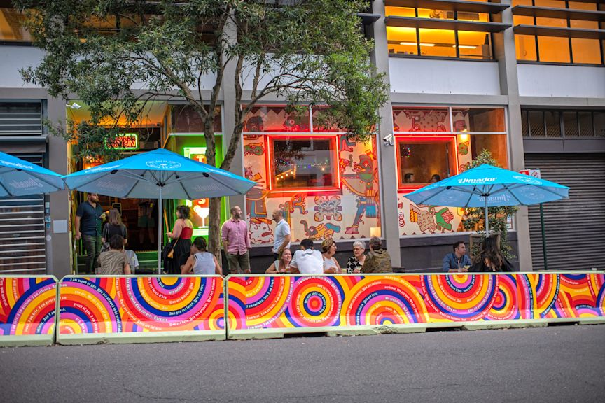 People sit and talk at outdoor tables under blue umbrellas outside a colorful, mural-covered restaurant on a city street.