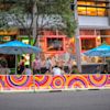 People sit and talk at outdoor tables under blue umbrellas outside a colorful, mural-covered restaurant on a city street.