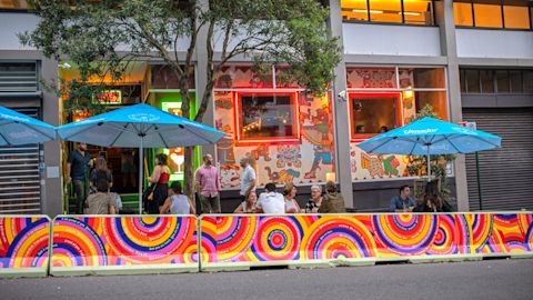 People sit and talk at outdoor tables under blue umbrellas outside a colorful, mural-covered restaurant on a city street.