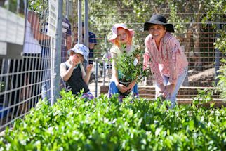 A woman and two children, all wearing sun hats, smile while holding plants in a garden near a wire fence.