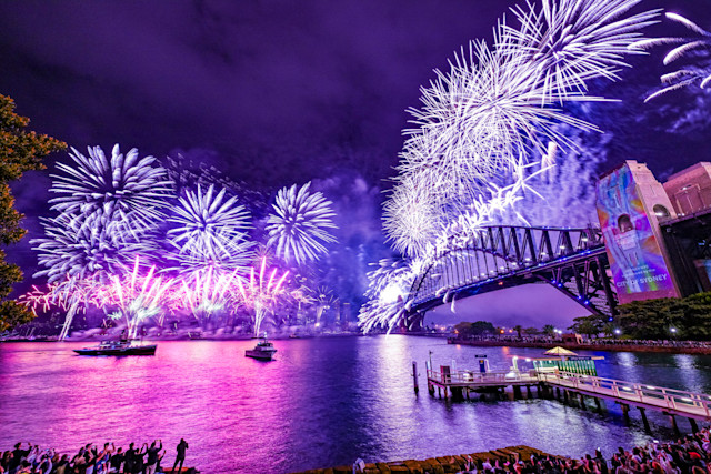New Year's fireworks over the harbour with the Sydney Harbour Bridge as the centrepiece. Spectators watch from the shore and boats. Bright, colorful fireworks light up the sky.