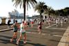 Runners participate in a marathon along a waterfront road near the Sydney Opera House and palm trees on a sunny day.
