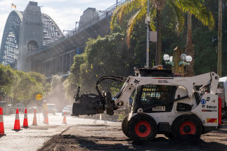 A white Bobcat skid-steer loader with a backhoe attachment works on a road construction site. Orange traffic cones outline the work area. The Sydney Harbour Bridge is in the background on a bright sunlit day.