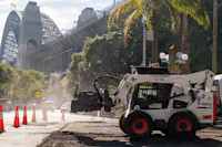 A white Bobcat skid-steer loader with a backhoe attachment works on a road construction site. Orange traffic cones outline the work area. The Sydney Harbour Bridge is in the background on a bright sunlit day.