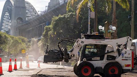 A white Bobcat skid-steer loader with a backhoe attachment works on a road construction site. Orange traffic cones outline the work area. The Sydney Harbour Bridge is in the background on a bright sunlit day.