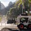 A white Bobcat skid-steer loader with a backhoe attachment works on a road construction site. Orange traffic cones outline the work area. The Sydney Harbour Bridge is in the background on a bright sunlit day.